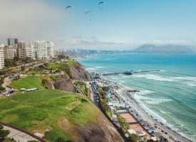 Stadtansicht entlang der Küste von Lima in Peru mit Hochhäusern, Klippen, grünen Parkflächen, einem Pier, der ins Meer ragt, und drei Gleitschirmfliegern am Himmel.