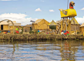 Ein Dorf am See mit traditionellen Strohdächern und großen gelben vogelförmigen Strukturen aus Schilf, mit Menschen, die nahe am Wasser täglichen Aktivitäten nachgehen, unter einem teils bewölkten Himmel.