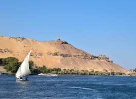 A sailboat gliding on the river in front of a hill with ancient ruins and a small structure at the top.