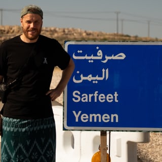 A man stands next to a blue road sign showing directions and distances to Sarfeet and Yemen in both Arabic and English, set in a dry, rocky landscape under a clear sky.