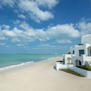 Weiße moderne Gebäude mit Treppen und Terrassen an einem Sandstrand mit ruhigem türkisfarbenem Meer und teilweise bewölktem Himmel.