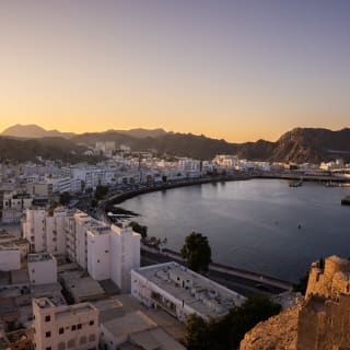 Sunset view of Muscat's coastal cityscape with buildings curving around a large bay, mountains in the background, and a clear sky.