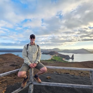 Ein Mann mit Kappe, Sonnenbrille und Wanderkleidung sitzt auf einem Holzgeländer mit Panoramablick auf vulkanische Inseln und Ozean im Hintergrund unter einem teils bewölkten Himmel.