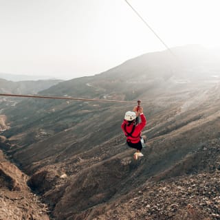 A person wearing a helmet and harness glides on a zip line over rugged terrain in Ras Al Khaimah, with mountains in the background.