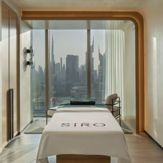 Ein modernes Wellness- oder Behandlungszimmer mit einer Massagebank, großen Fenstern mit Stadt-Skyline und minimalistischer Dekoration.