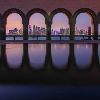 View of Doha skyline at dusk framed by a series of stone arches reflected in a calm pool of water, with a person standing near the right side arch.