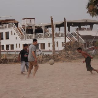 Gruppe von Menschen spielt barfuß Fußball auf einem sandigen Feld in der Nähe von Palmen und einem großen weißen Gebäude in Fayoum, Ägypten.