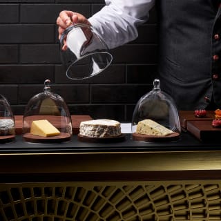 A waiter presents a selection of cheese under glass covers on a trolley, with another tray of desserts on the side.