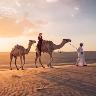Eine Person führt zwei Kamele durch eine Wüste bei Sonnenuntergang, mit einer weiten Sandlandschaft und einem bunten Himmel in Oman.
