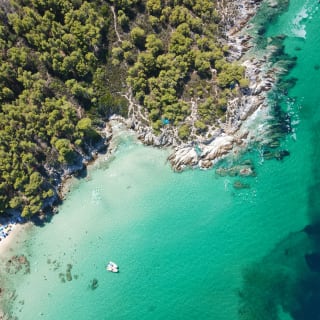 Luftaufnahme einer bewaldeten Küstenlinie mit klarem türkisfarbenem Wasser und einem kleinen Sandstrand mit Sonnenschirmen, mit felsigen Uferbereichen.