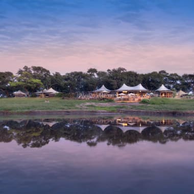 Somalisa Camp in Simbabwe liegt an einem Fluss, mit Zelten und Bäumen, die im Wasser gespiegelt werden.