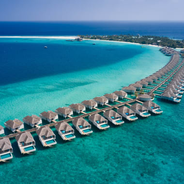 Aerial view of overwater bungalows extending from a tropical island into turquoise ocean waters under a clear blue sky.