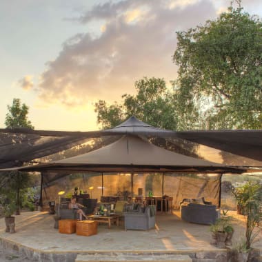 A luxurious outdoor lounge area under a large canopy with sofas, chairs, and tables, surrounded by trees during sunset.