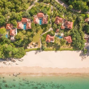 Luftaufnahme eines Strandresorts mit mehreren Villen, umgeben von üppigem Grün, neben einem Sandstrand und klarem türkisfarbenem Wasser.