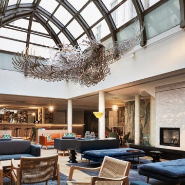 Geräumige moderne Hotellobby mit hoher Glasdecke, großer Metallvogelskulptur, gemütlichen blauen und orangen Sitzbereichen, Holzstühlen, Kamin und Bar im Hintergrund.