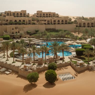 Ein luxuriöses Wüstenresort mit einem Pool, umgeben von Palmen und Sonnenliegen, in einer trockenen Wüstenumgebung.