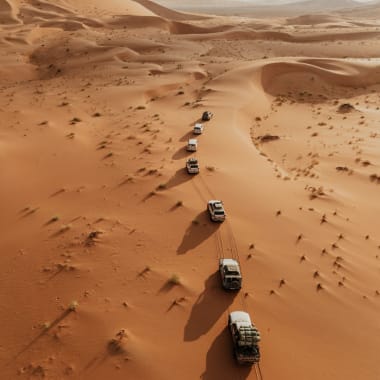 Sieben Geländewagen in einer Reihe fahren durch die Sanddünen der Wüste, mit langen Schatten, die von der Sonne auf der linken Seite an den Fahrzeugen geworfen werden.
