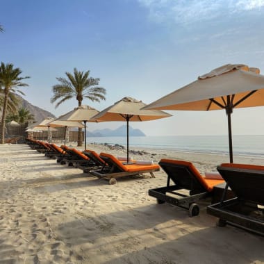 Eine Reihe leerer Liegestühle mit orangefarbenen Kissen und großen Sonnenschirmen am sandigen Strand, mit Palmen und Bergen im Hintergrund.