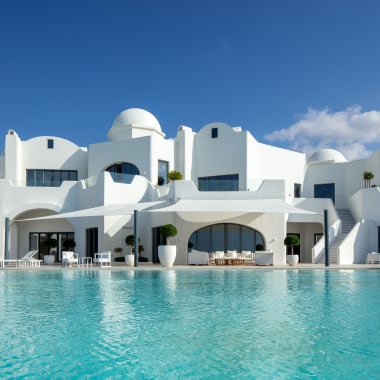 Ein luxuriöses weißes Hotel mit runder Architektur und großen Glasfenstern am Pool unter einem blauen Himmel.