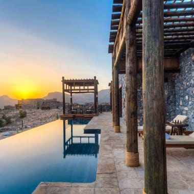 Luxury villa with a swimming pool overlooking a sunset in a mountainous area with stone walls and wooden pillars.