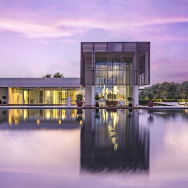 A modern resort with a central building reflected in a large, still pool, during sunset with purple and pink sky.