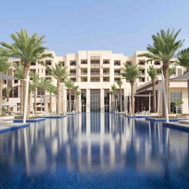 Luxuriöses Hotelkomplex mit großem Pool, umgeben von Palmen und modernen Gebäuden unter klarem blauem Himmel.