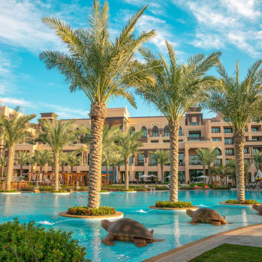 Luxuriöses Resort mit großem Swimmingpool, umgeben von Palmen und Schildkrötenstatuen, mit Gebäuden im Hintergrund unter einem leicht bewölkten Himmel.
