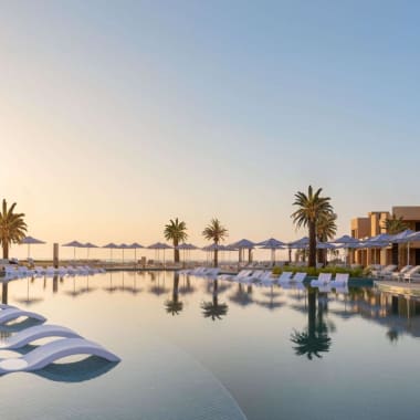 Ein luxuriöser Hotelpoolbereich bei Sonnenuntergang mit Liegestühlen, Sonnenschirmen und Palmen, die sich im Wasser spiegeln.