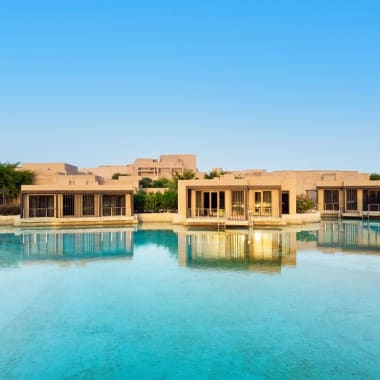 Luxury resort buildings reflected in a calm water body against a clear blue sky.