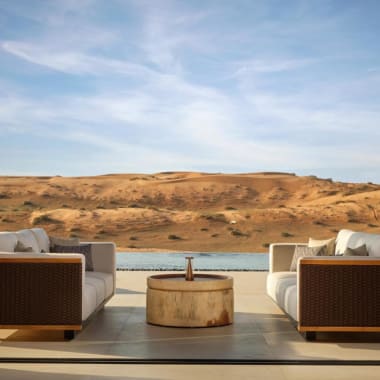 Zwei Sofas, die sich gegenüberstehen, mit einem kleinen runden Tisch dazwischen auf einer Terrasse mit Blick auf Sanddünen und einen Fluss unter einem teilweise bewölkten Himmel.