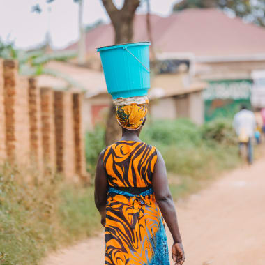 A dark-skinned woman in traditional dress carrying a bucket on her head