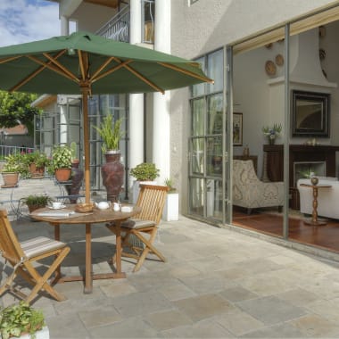 Eine sonnige Terrasse mit einem runden Holztisch, vier Holzstühlen und einem großen grünen Sonnenschirm vor einem Haus mit Glastüren, die in ein Wohnzimmer führen.