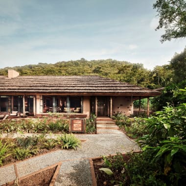 A single-story lodge with large windows, surrounded by lush greenery and a garden path leading up to it, set against a backdrop of forested hills.