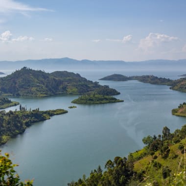 A panoramic view of Lake Kivu surrounded by hills and lush greenery under a partly cloudy sky.