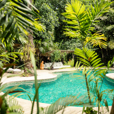 Curved turquoise swimming pool surrounded by lush tropical plants and trees, with white lounge chairs and a cozy seating area in the background.