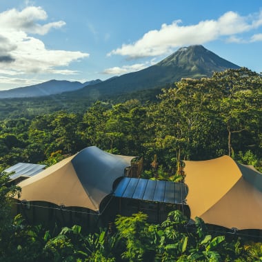 Luftaufnahme des Nayara Tented Camp, eingebettet in einen üppigen tropischen Wald mit einem markanten Vulkan im Hintergrund unter einem teilweise bewölkten Himmel.