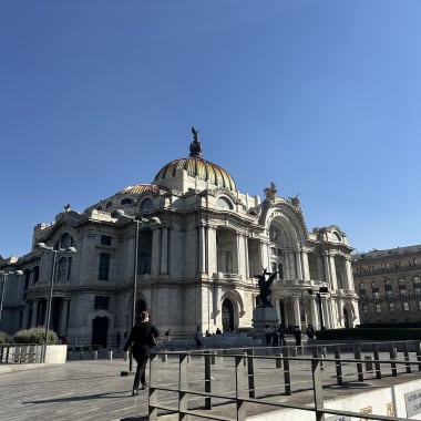 Das Palacio de Bellas Artes in Mexiko-Stadt unter klarem blauem Himmel, mit einigen Menschen, die in der Nähe auf einem gepflasterten Platz gehen.
