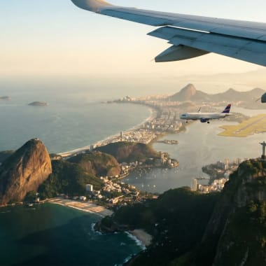 Luftaufnahme der Küste von Rio de Janeiro mit dem Zuckerhut, der Christus-Erlöser-Statue und einem Flugzeug, das nahe der Küste unter einem Flugzeugflügel fliegt.