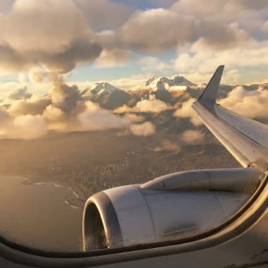 Blick aus dem Flugzeugfenster auf das Flugzeugflügel und Triebwerk mit schneebedeckten Bergen und Wolken in der Ferne bei Sonnenuntergang.