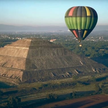 Ein bunter Heißluftballon schwebt neben der antiken Sonnenpyramide von Teotihuacan, Mexiko, umgeben von Grünflächen und entfernten Bergen.