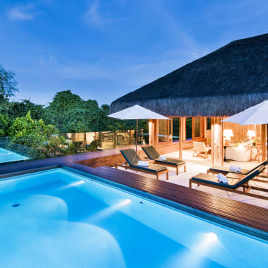 A luxurious outdoor pool area at a tropical resort with sun loungers and umbrellas, adjacent to a thatched-roof villa.