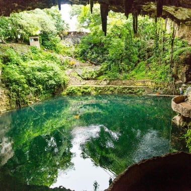 Innerhalb einer Höhle mit einem türkisfarbenen Wasserbecken, umgeben von dichtem Grün und felsigen Wänden, mit Stalaktiten an der Decke.