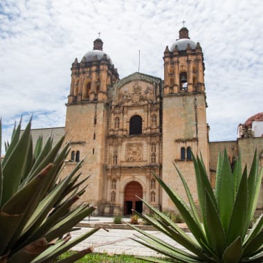 Historische Steinkirche mit zwei Türmen in Oaxaca, eingefasst von Agavenpflanzen unter einem teilweise bewölkten Himmel.