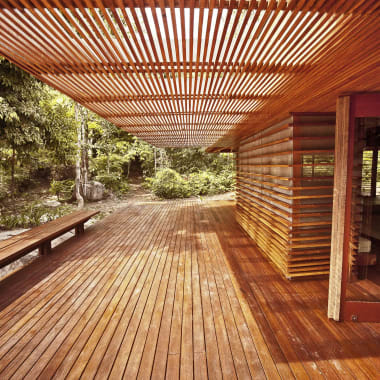 Spacious wooden deck with slatted wood ceiling and bench, next to a modern lodge surrounded by lush greenery.