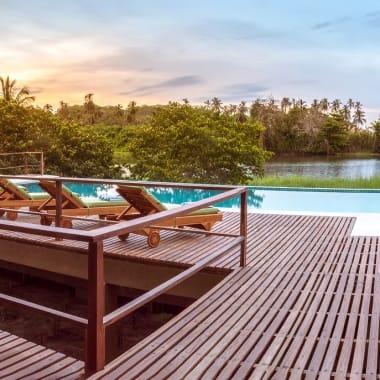 Holzdeck mit Liegestühlen um einen Infinity-Pool mit Blick auf üppiges Grün und ein ruhiges Meer unter einem teils bewölkten Himmel bei Sonnenuntergang.