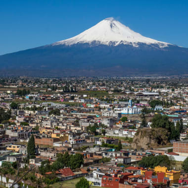 Luftaufnahme der Stadt Puebla mit zahlreichen Gebäuden und Straßen im Vordergrund, vor dem Hintergrund eines großen schneebedeckten Vulkans unter klarem blauem Himmel.