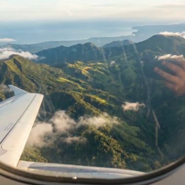 Blick aus dem Flugzeugfenster auf grüne Berge und Wolken über einer Küstenlandschaft bei Tageslicht.