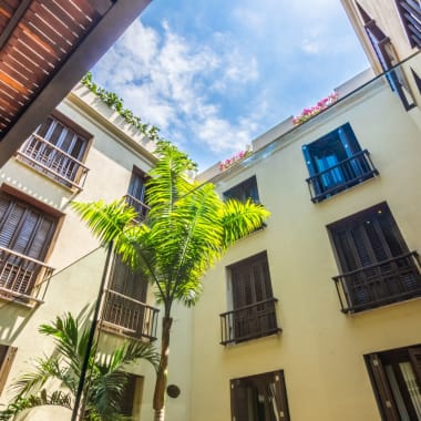 View from the courtyard of a hotel looking up at the sky, surrounded by cream-colored walls with black window shutters and balconies.