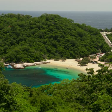 Abgeschiedene Strandbucht mit türkisfarbenem Wasser, umgeben von üppigen grünen Hügeln und dichtem Wald in Huatulco, Mexiko.
