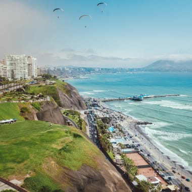 Stadtansicht entlang der Küste von Lima in Peru mit Hochhäusern, Klippen, grünen Parkflächen, einem Pier, der ins Meer ragt, und drei Gleitschirmfliegern am Himmel.